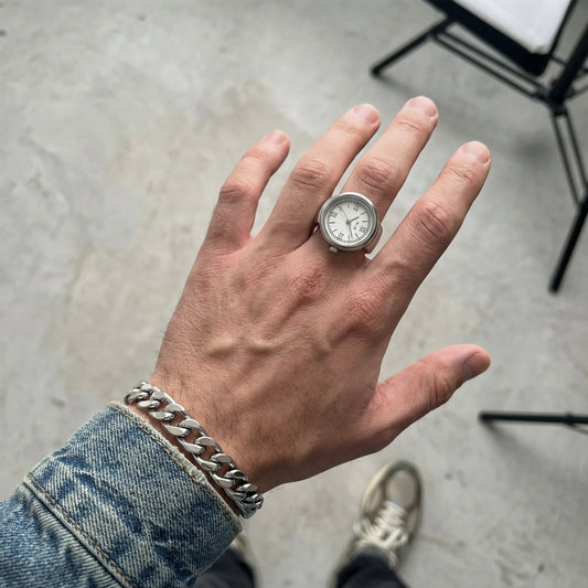 Hand wearing a silver ring with a watch face on a concrete floor.