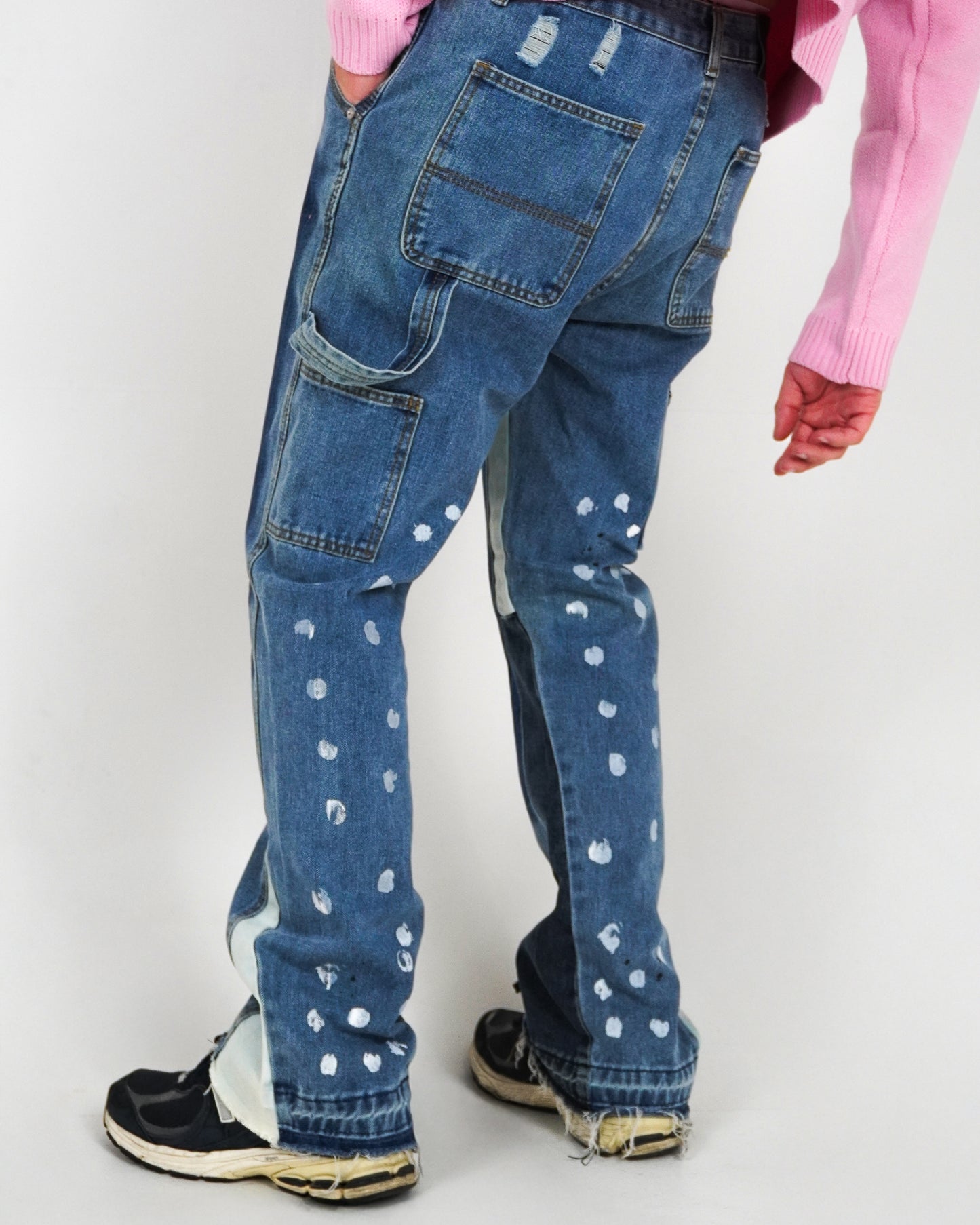 Person wearing blue jeans with paint splatters and a pink sweater, standing on a plain background.