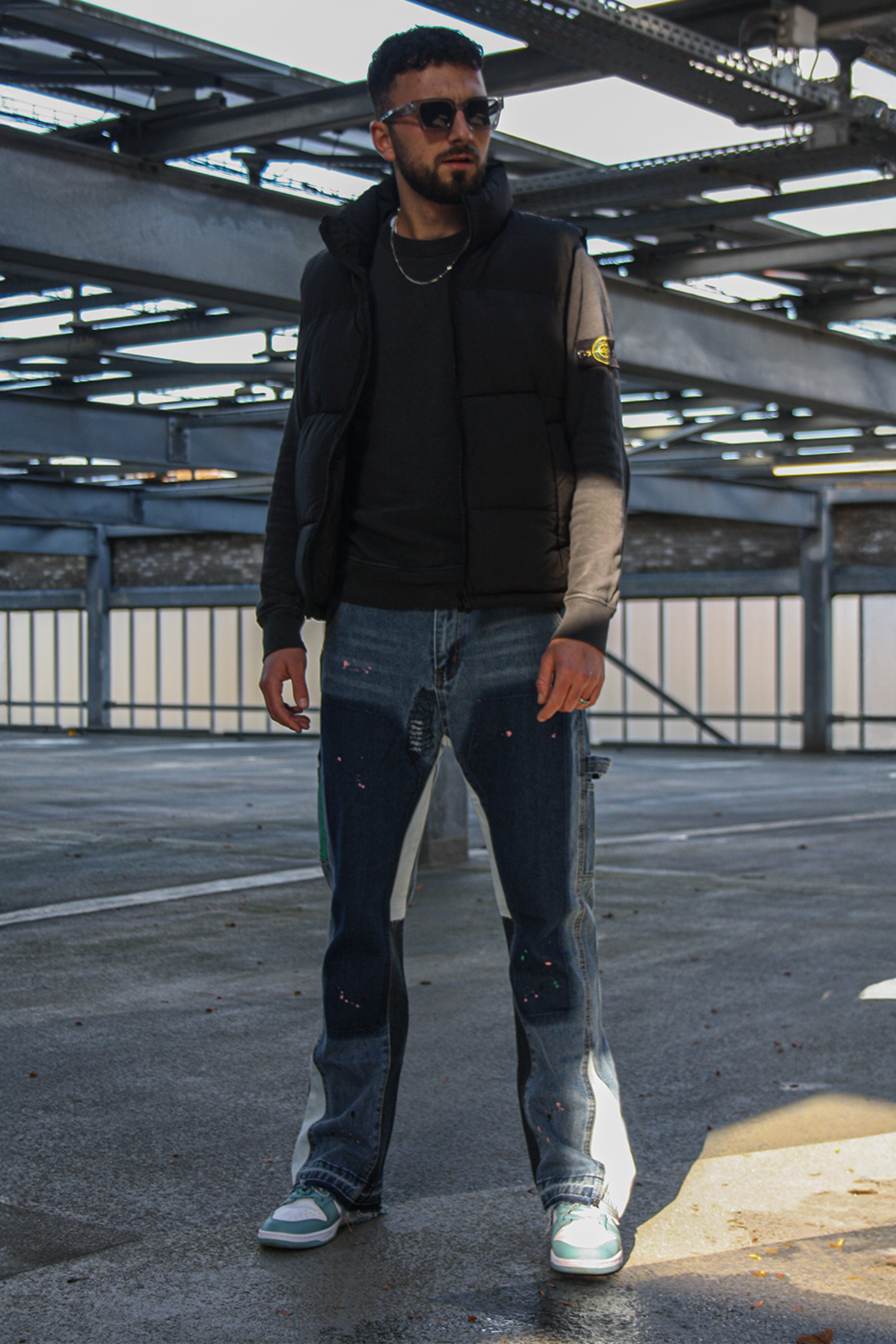 Man wearing a black jacket, black shirt, and blue jeans with white sneakers on an industrial rooftop.
