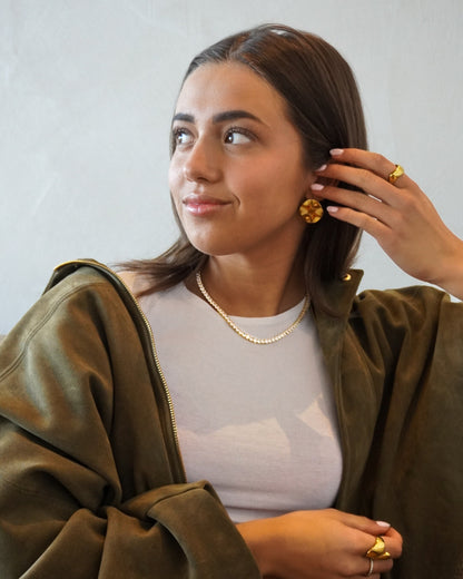 Person with brown hair wearing a white top and green jacket, holding hair and wearing gold earrings.