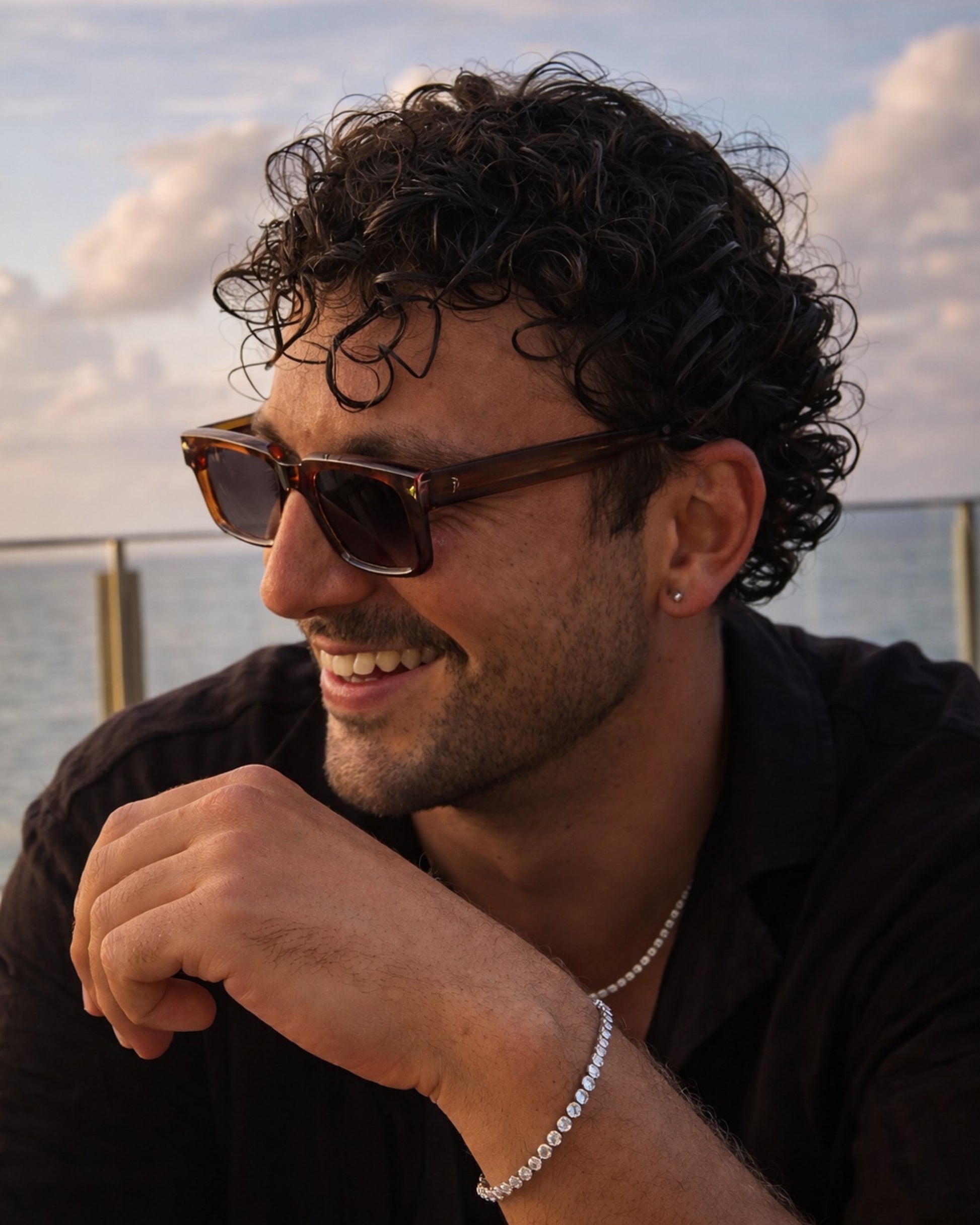 Person with curly hair wearing sunglasses and jewelry, smiling by the water.