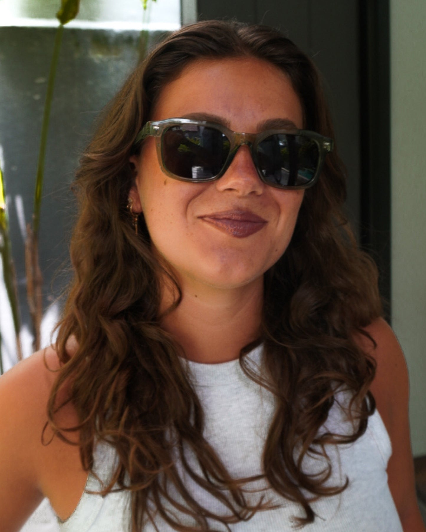 Person with wavy hair wearing sunglasses and a light tank top, smiling outdoors.