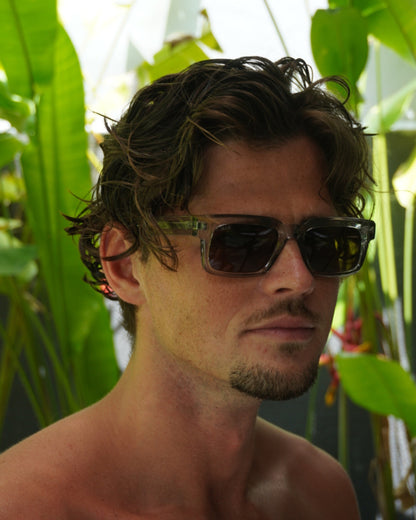 Person with short curly hair wearing sunglasses, surrounded by green plants.