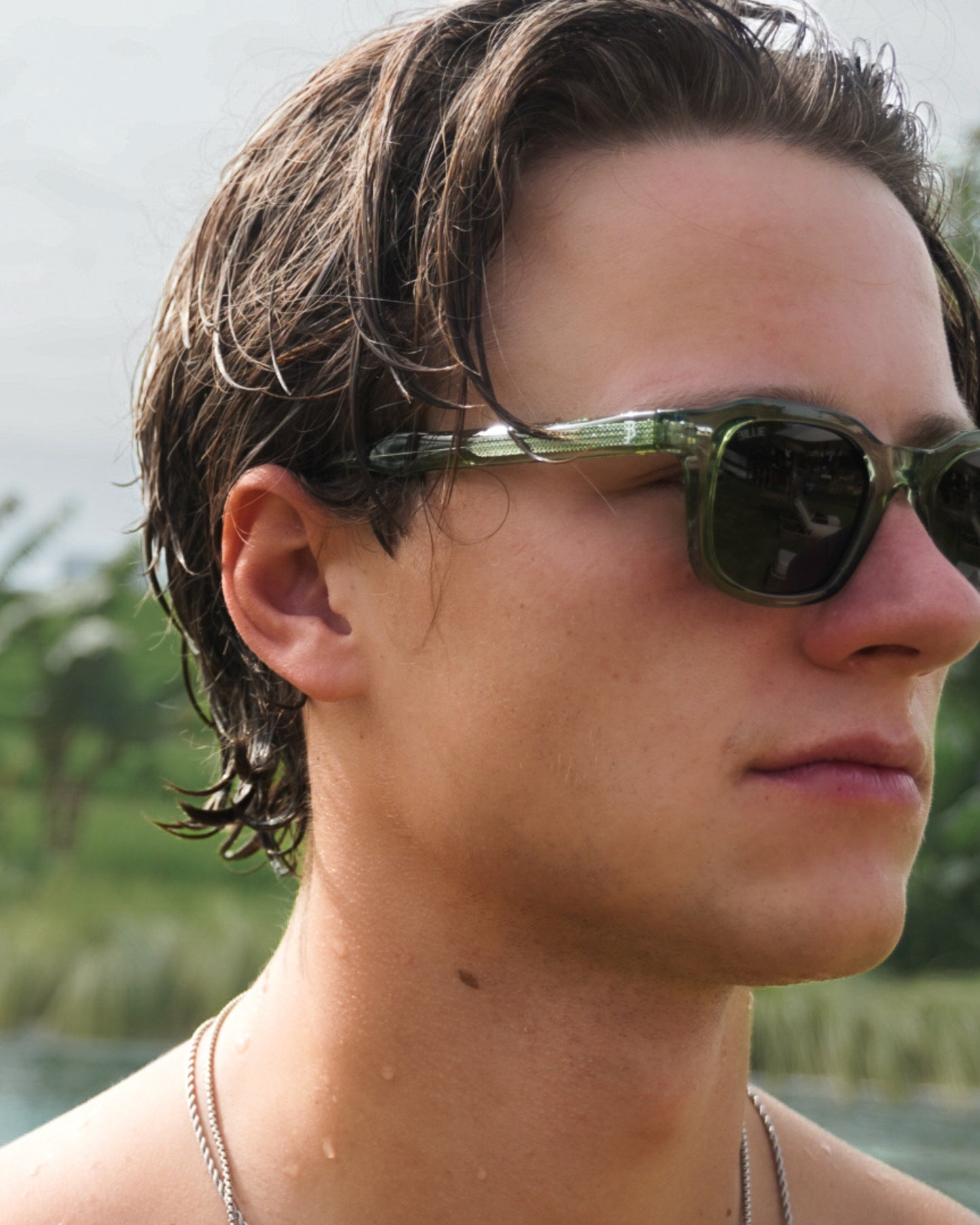 Close-up of a person with wet hair wearing green sunglasses, outdoors with greenery in the background.