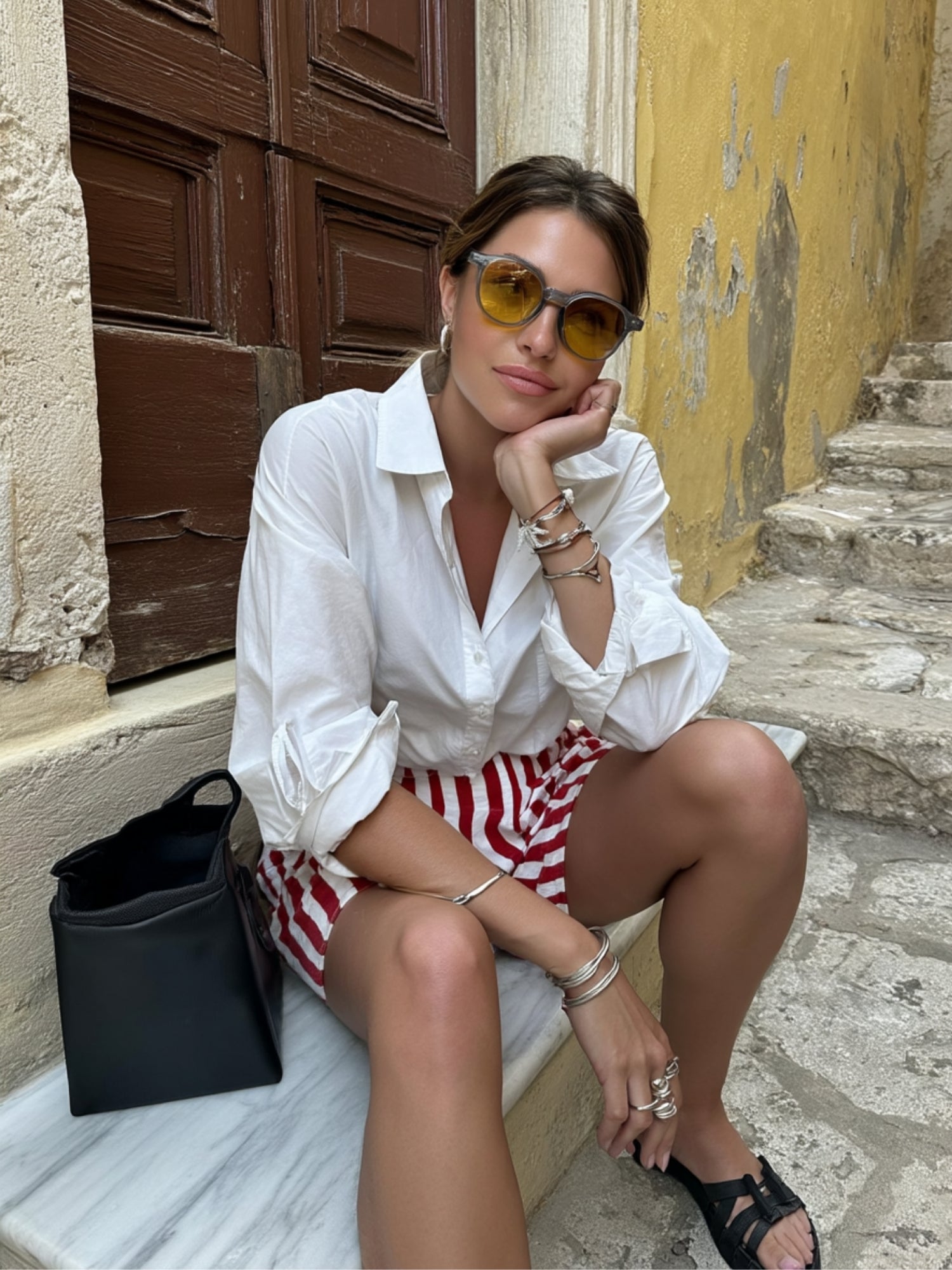 Woman sitting on steps wearing a white shirt and red and white striped shorts, with a black handbag.