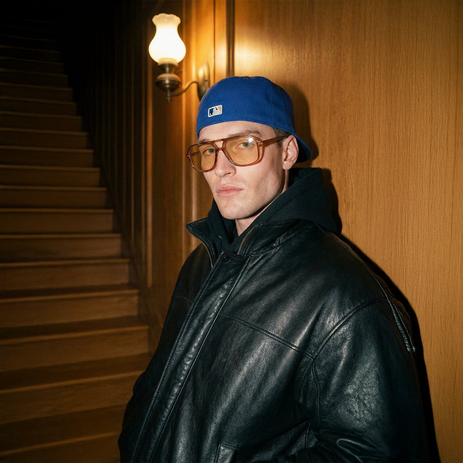 Man met een blauwe pet en een zwart leren jack, staande tegen een houten muur.