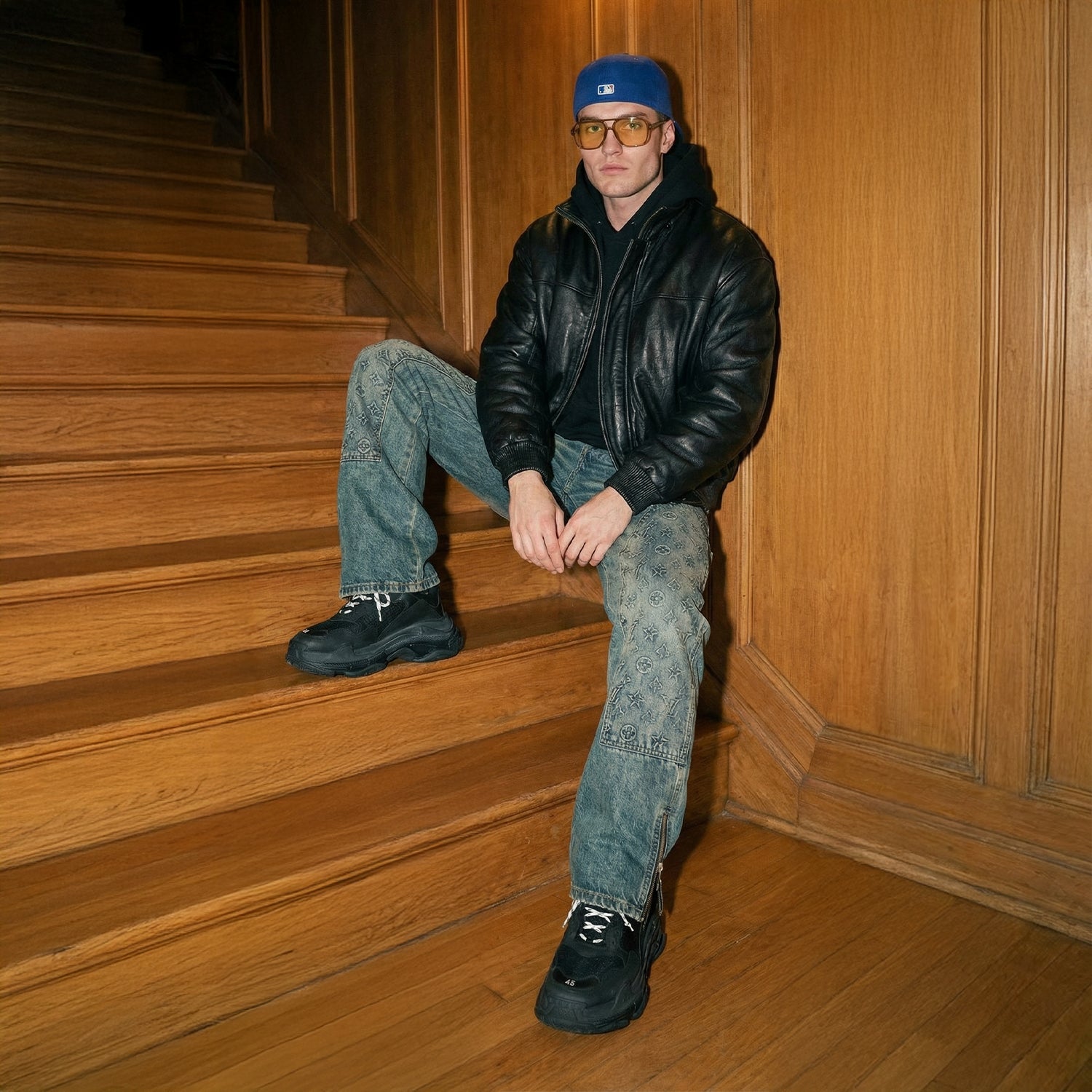 Persoon zittend op houten trap, gekleed in een zwart jasje, blauwe spijkerbroek en een blauwe pet.