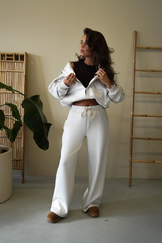 Woman in a white outfit standing in a minimalistic room with a plant and wooden shelves.