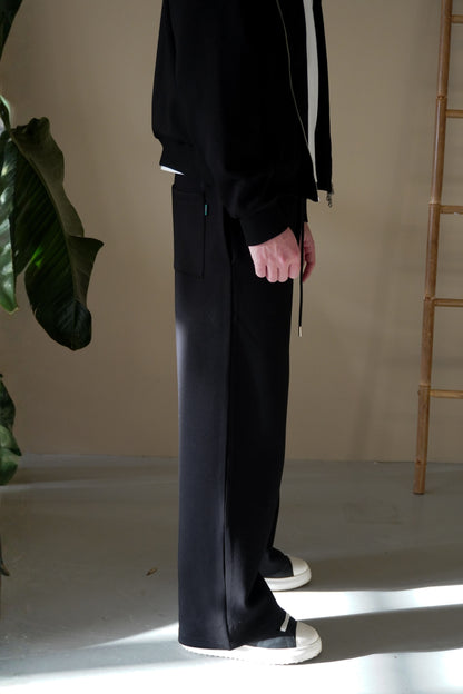 Person wearing black pants and a black jacket in a minimal indoor setting.