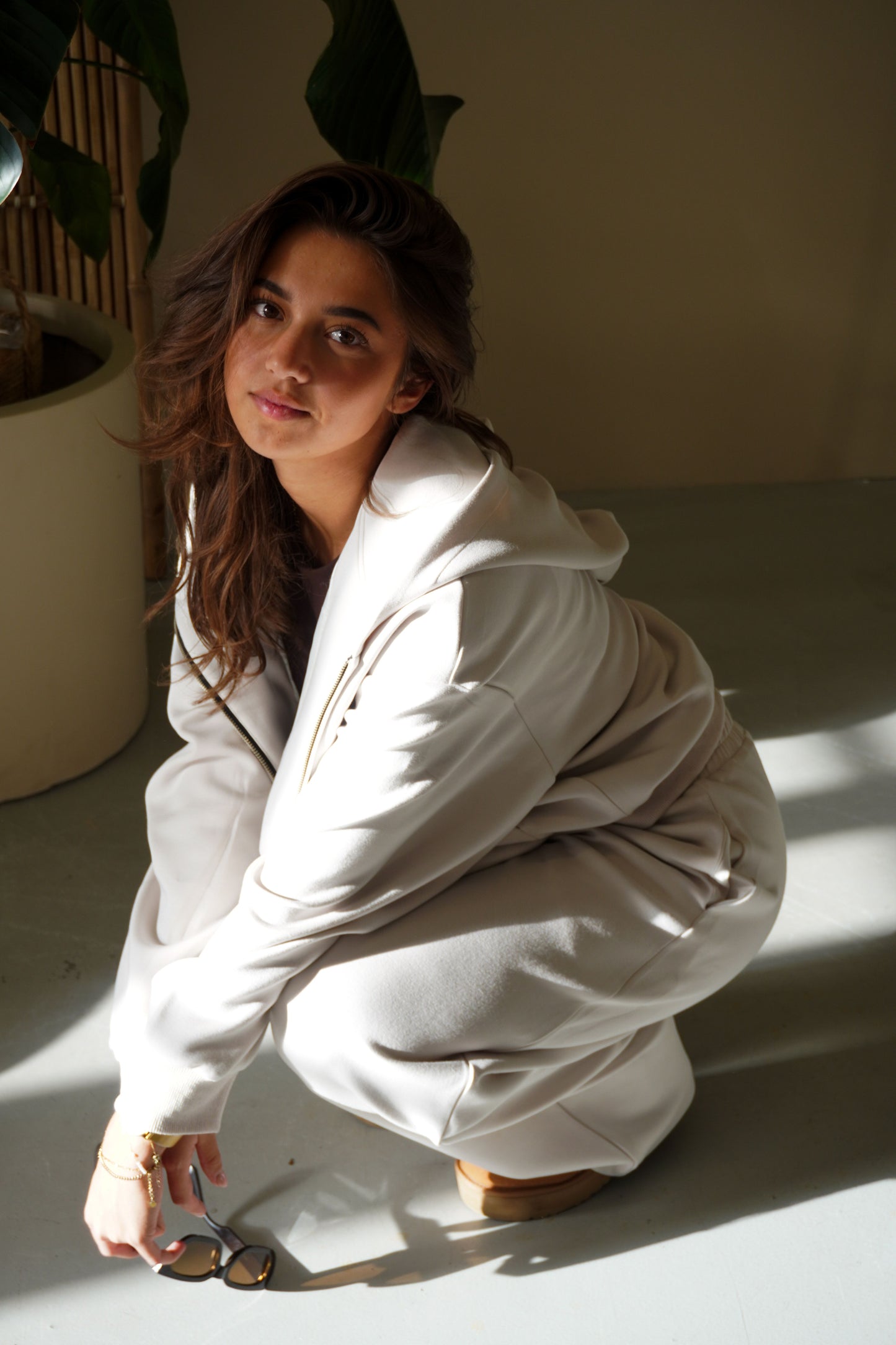Woman in a white outfit squatting on a light surface with a plant in the background