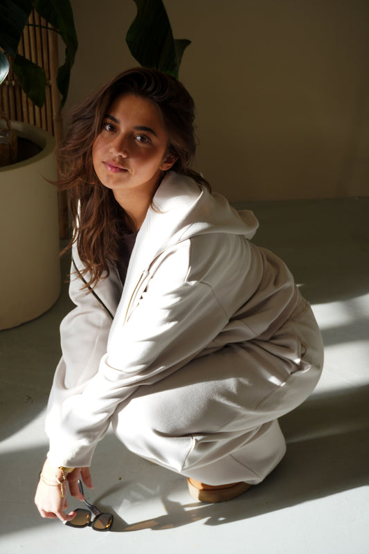Woman in a white outfit squatting on a light surface with a plant in the background