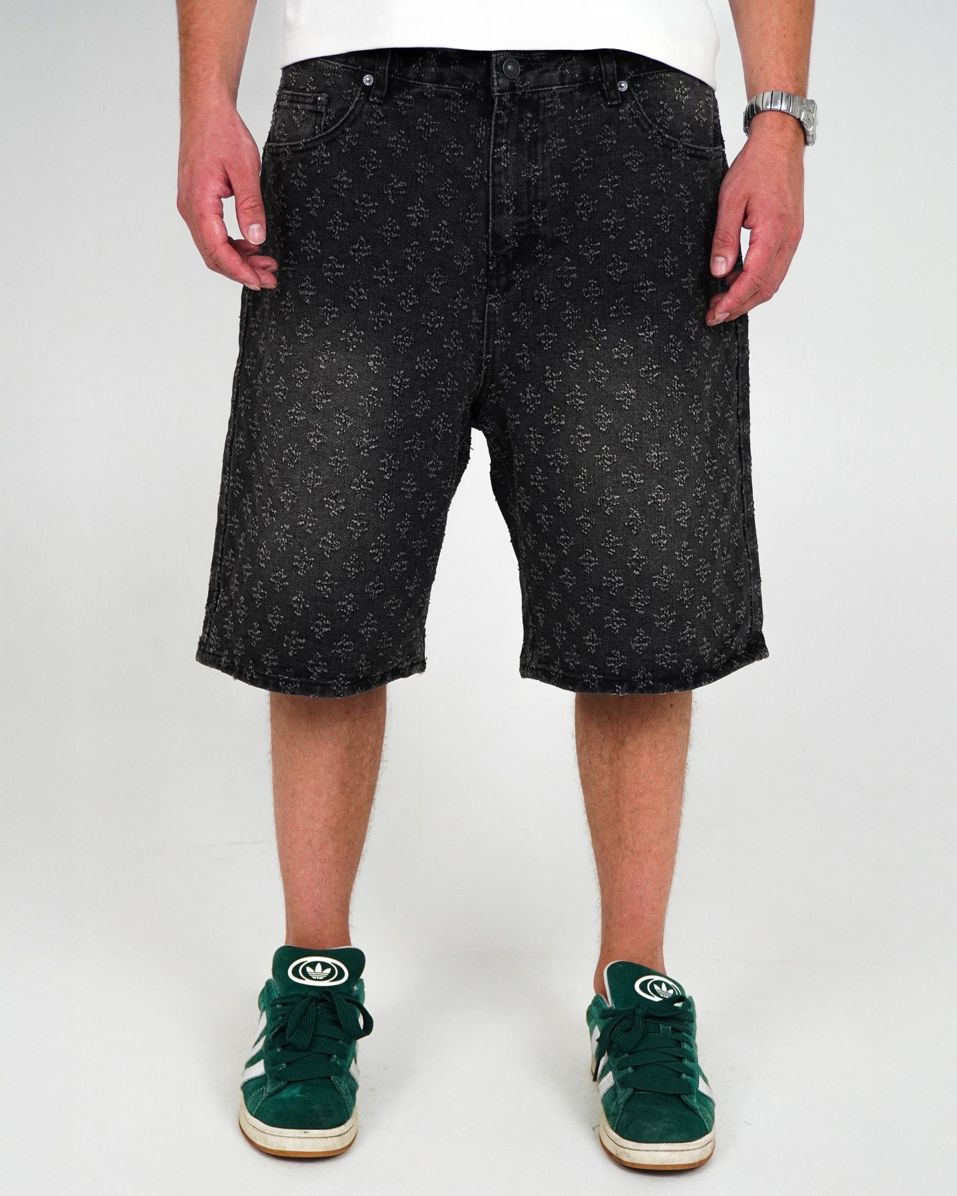 Person wearing black patterned shorts and green sneakers, standing on a plain background.