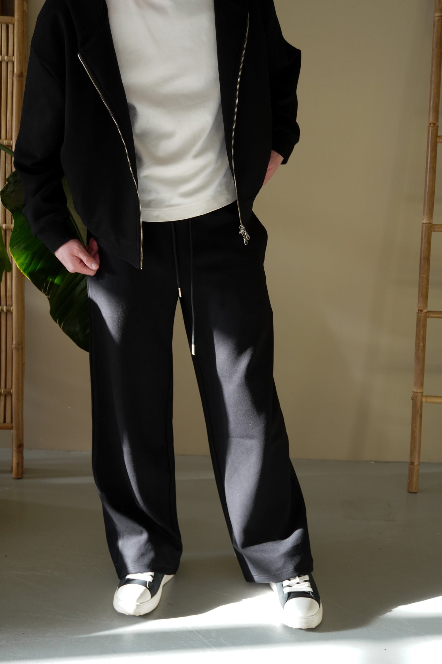 Person wearing a black zip up, white shirt, and black pants standing against a neutral background.