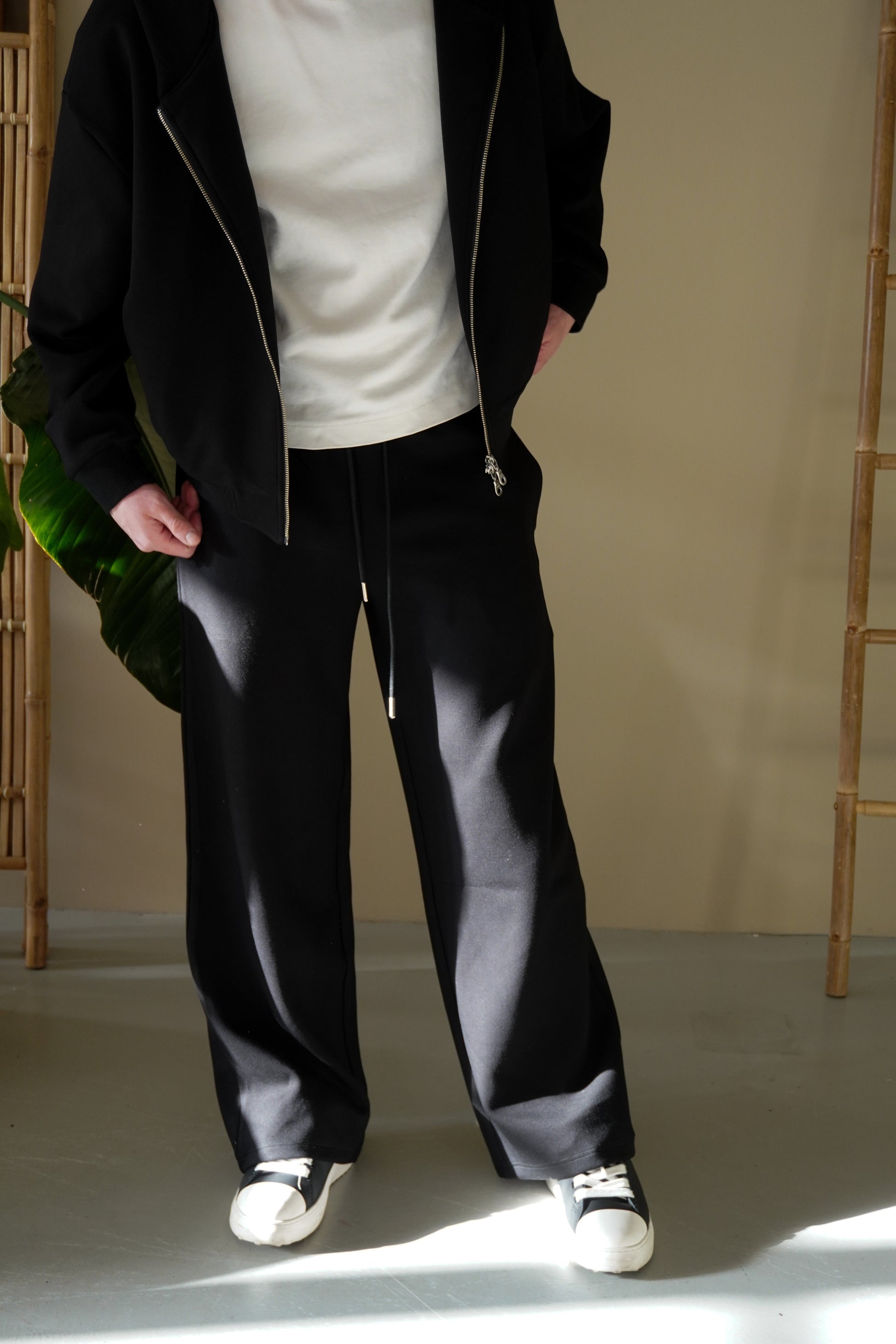 Person wearing a black zip up, white shirt, and black pants standing against a neutral background.