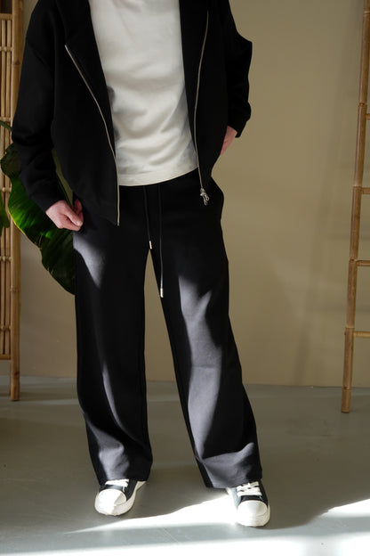 Person wearing a black zip up, white shirt, and black pants standing against a neutral background.