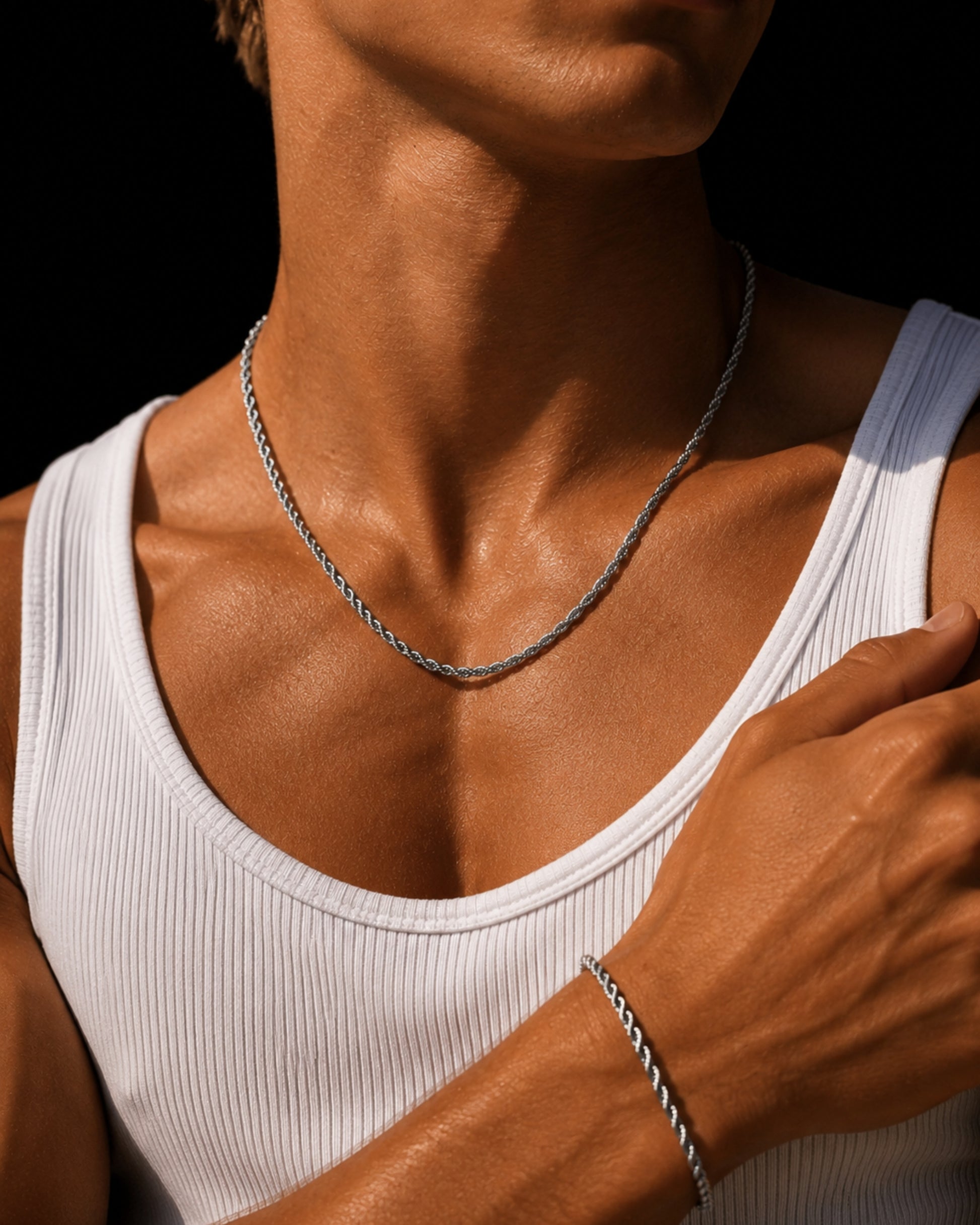 Close-up of a person's neck and arm wearing a silver chain and bracelet, with a white tank top.
