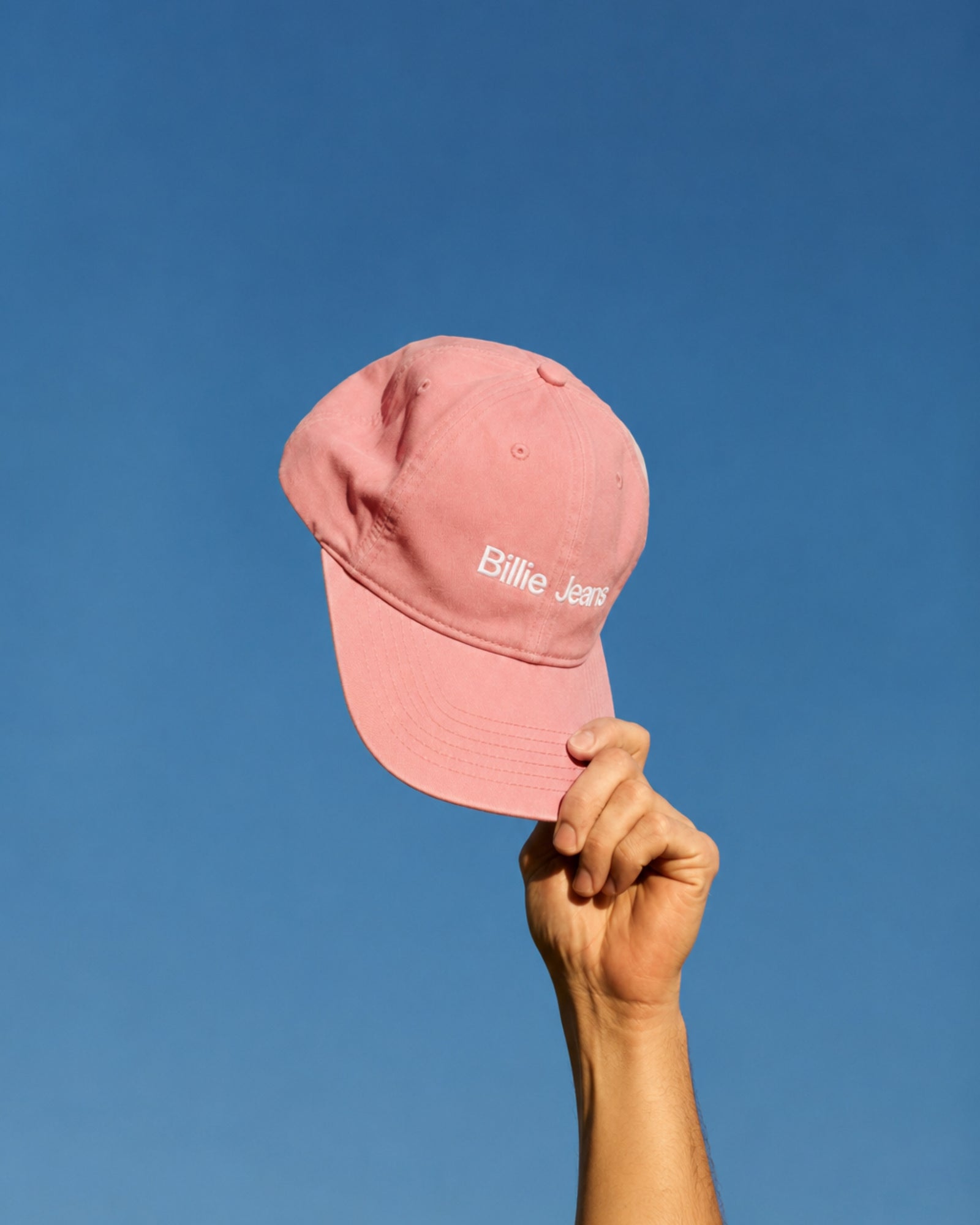 A hand holding a pink cap with the text 'Billie Jeans' against a blue sky.