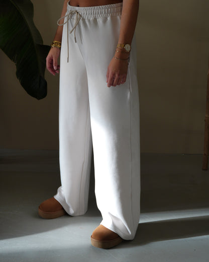Person wearing white wide-leg pants and brown shoes, standing on a light floor with a plant in the background.
