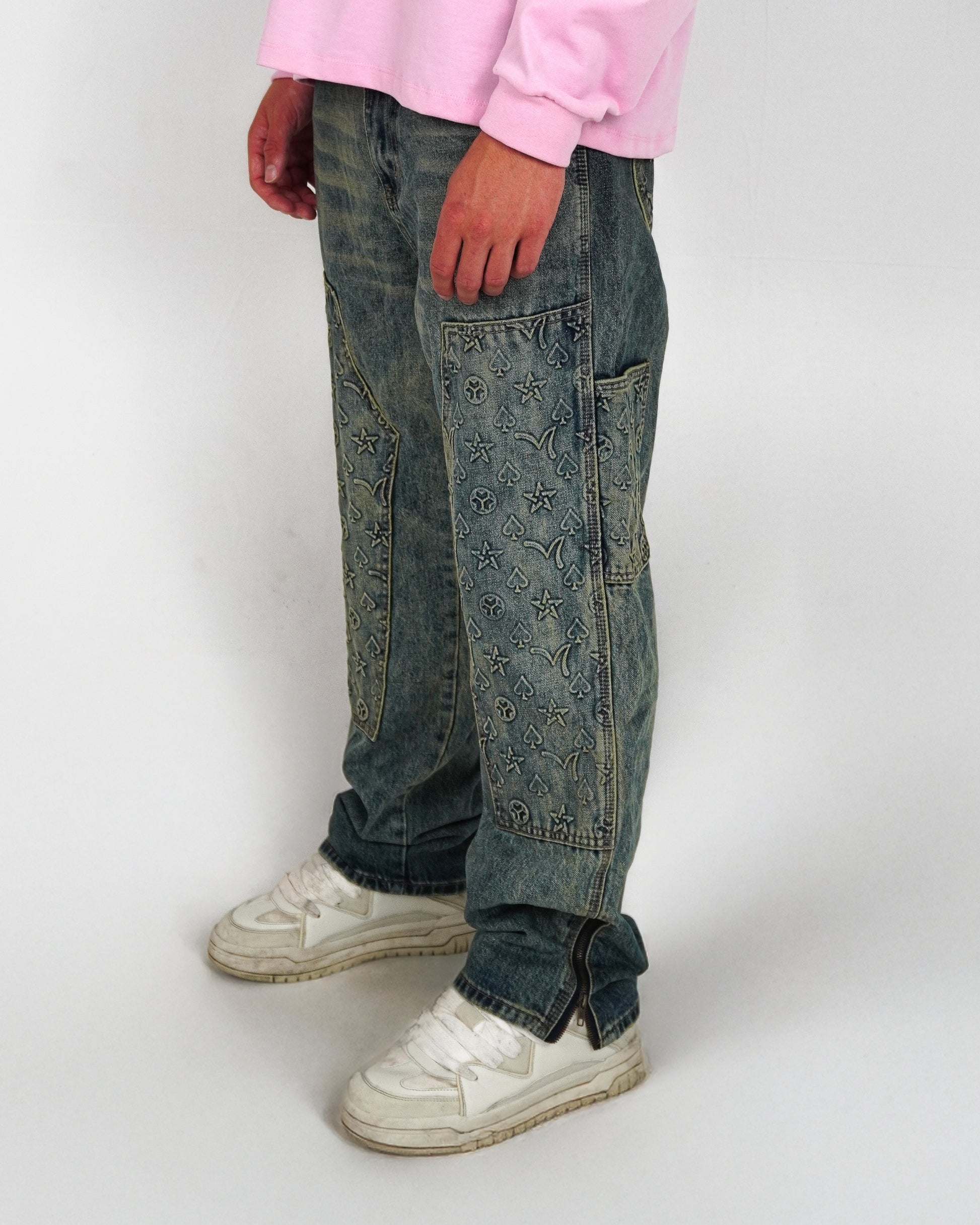 Person wearing blue jeans with decorative patterns and white sneakers, standing on a plain background.