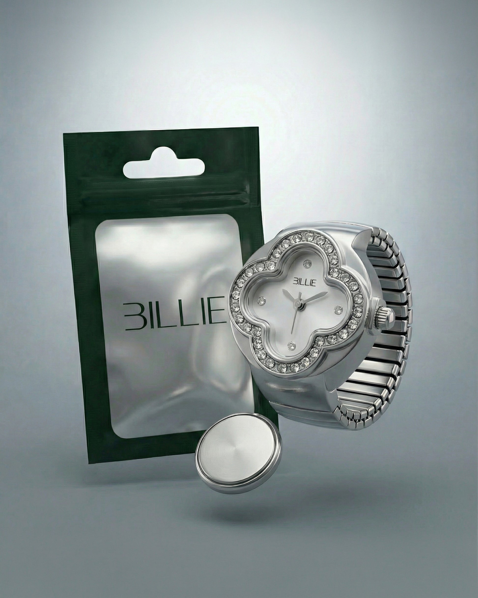 Silver watch with a floral face and crystals, next to a green packaging and a round silver object.