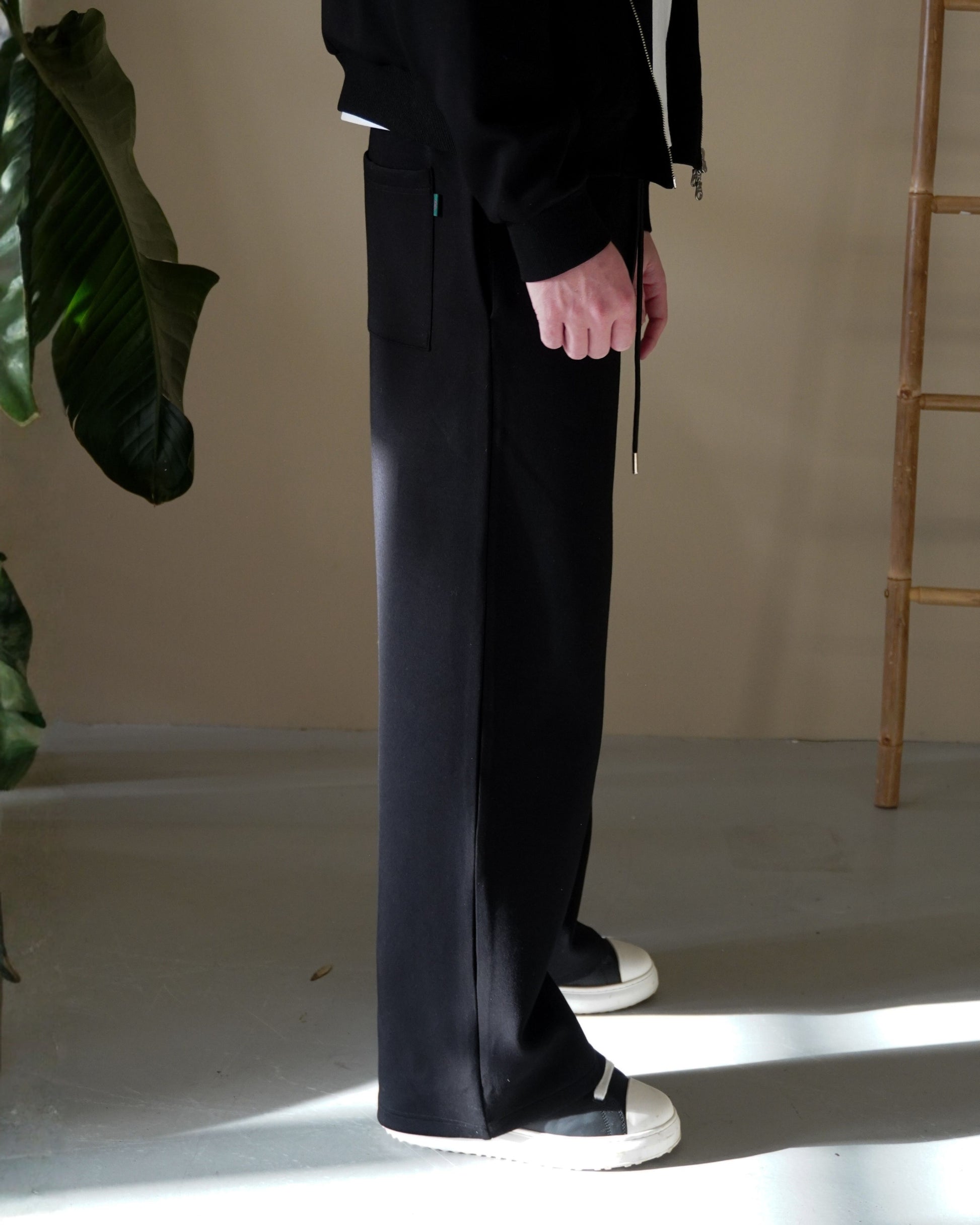 Person wearing black pants and white sneakers, standing beside a plant.