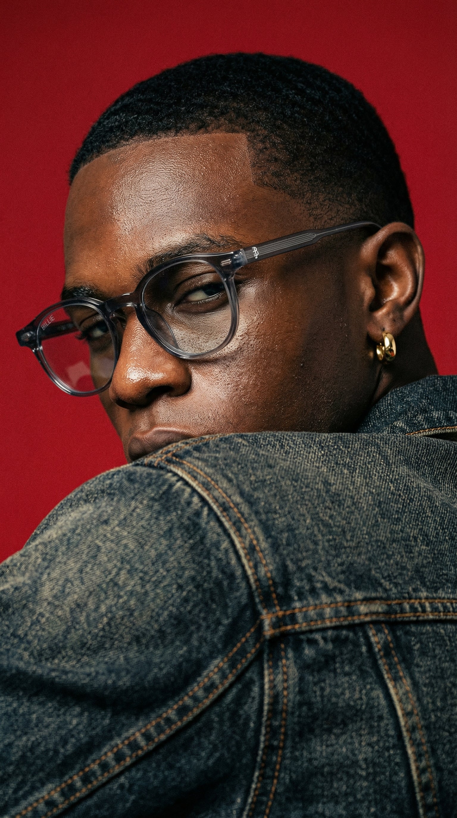 Person wearing glasses and a denim jacket against a red background