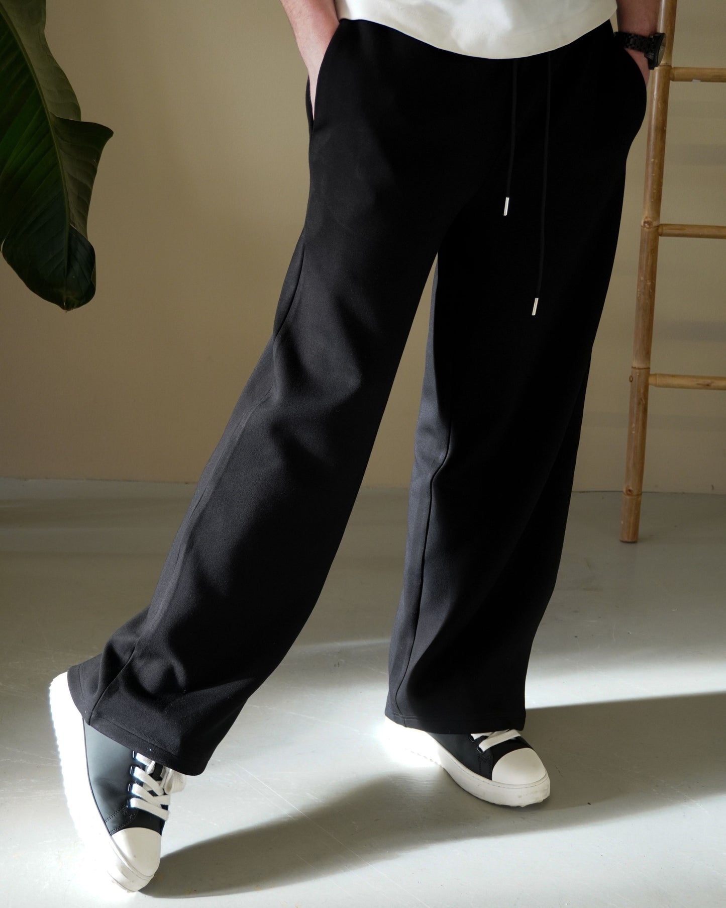 Person wearing black pants and black and white sneakers, standing indoors with a plant and wooden ladder.