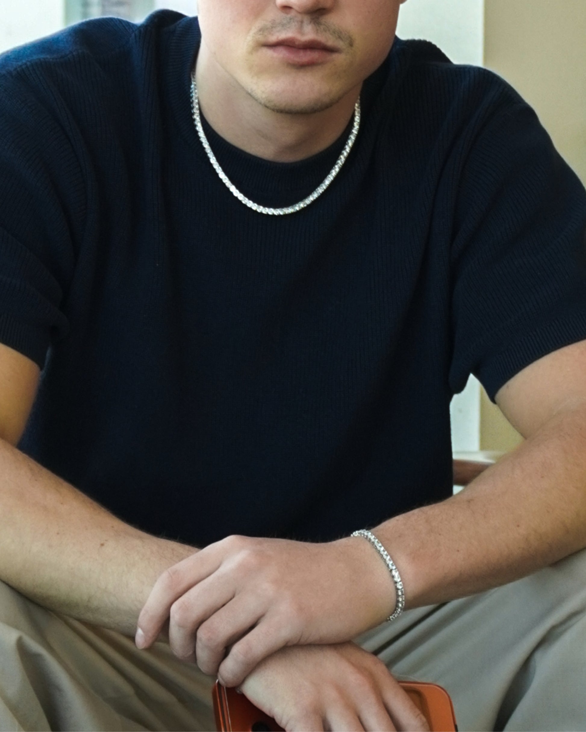 Person with short hair wearing a dark shirt and silver chain, sitting with hands on knees.
