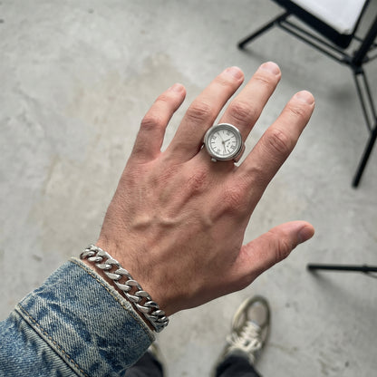 Hand wearing a silver ring with a watch face on a concrete floor.