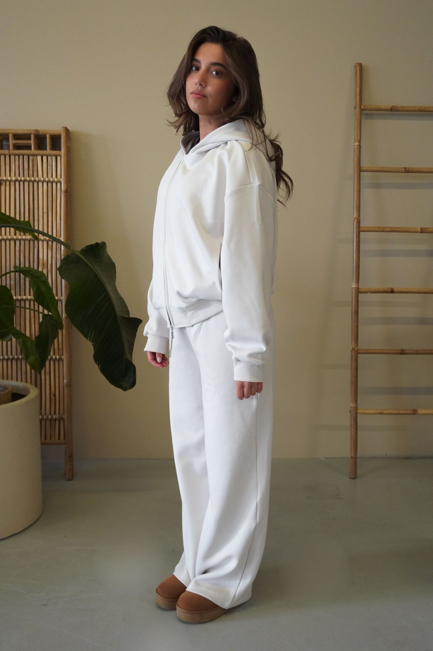 Woman wearing a white hoodie and pants standing in a minimalistic room with a plant and wooden ladder.