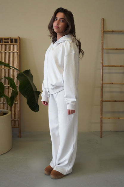 Woman wearing a white hoodie and pants standing in a minimalistic room with a plant and wooden ladder.