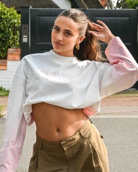 Person with long hair wearing a cropped white and pink top and khaki pants, posing outdoors.