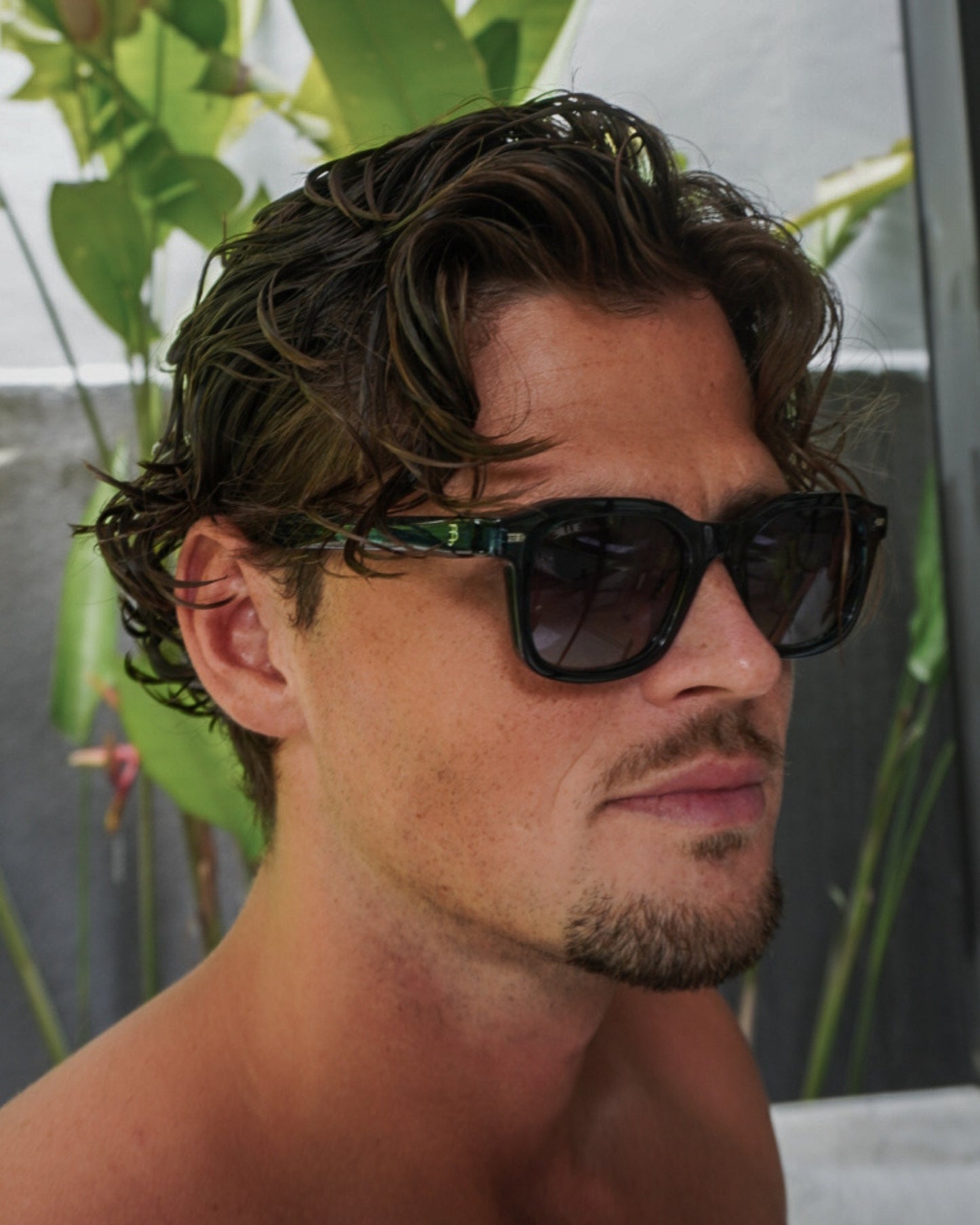 Person with curly hair wearing sunglasses, with green plants in the background.