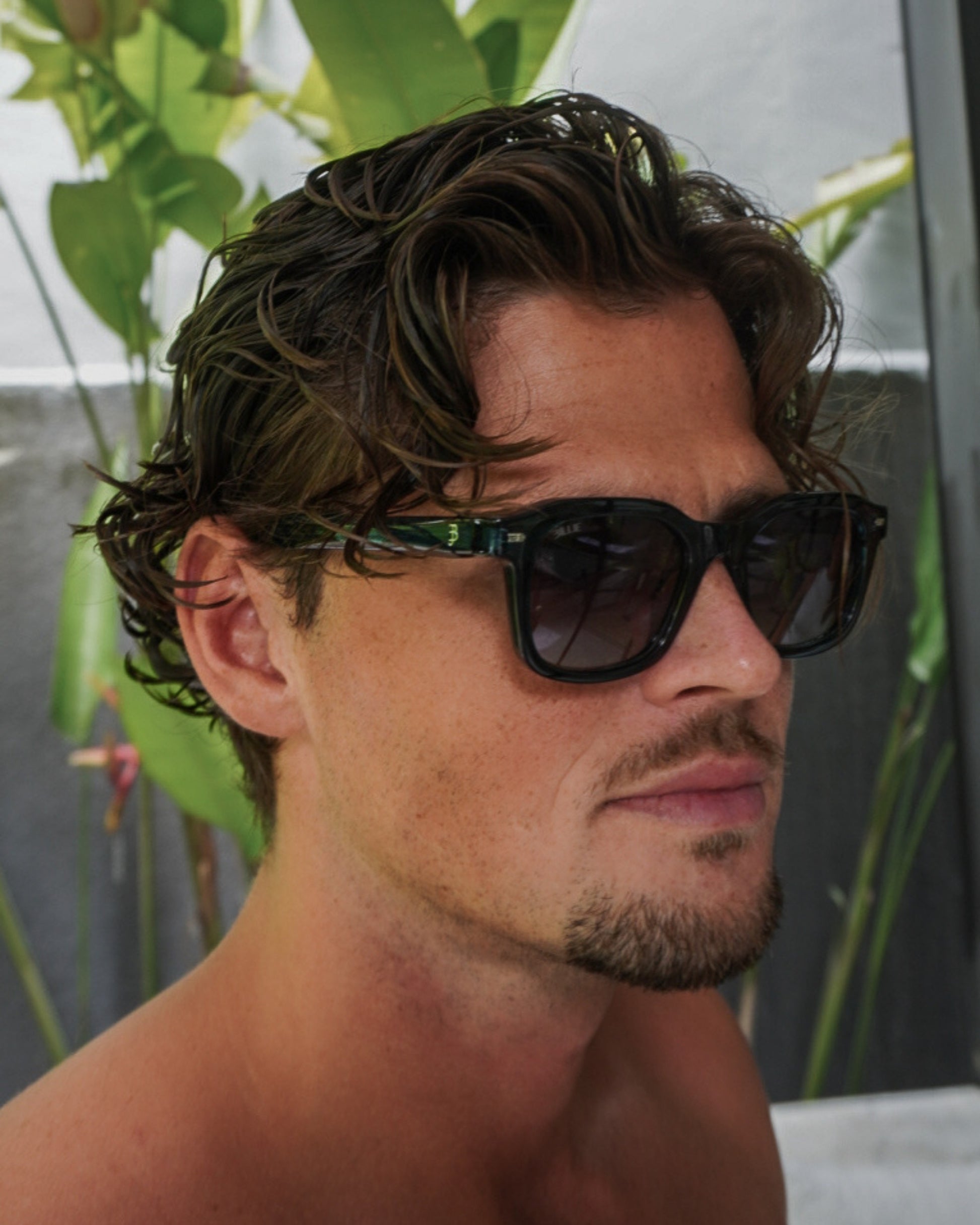 Person with curly hair wearing sunglasses, with green plants in the background.