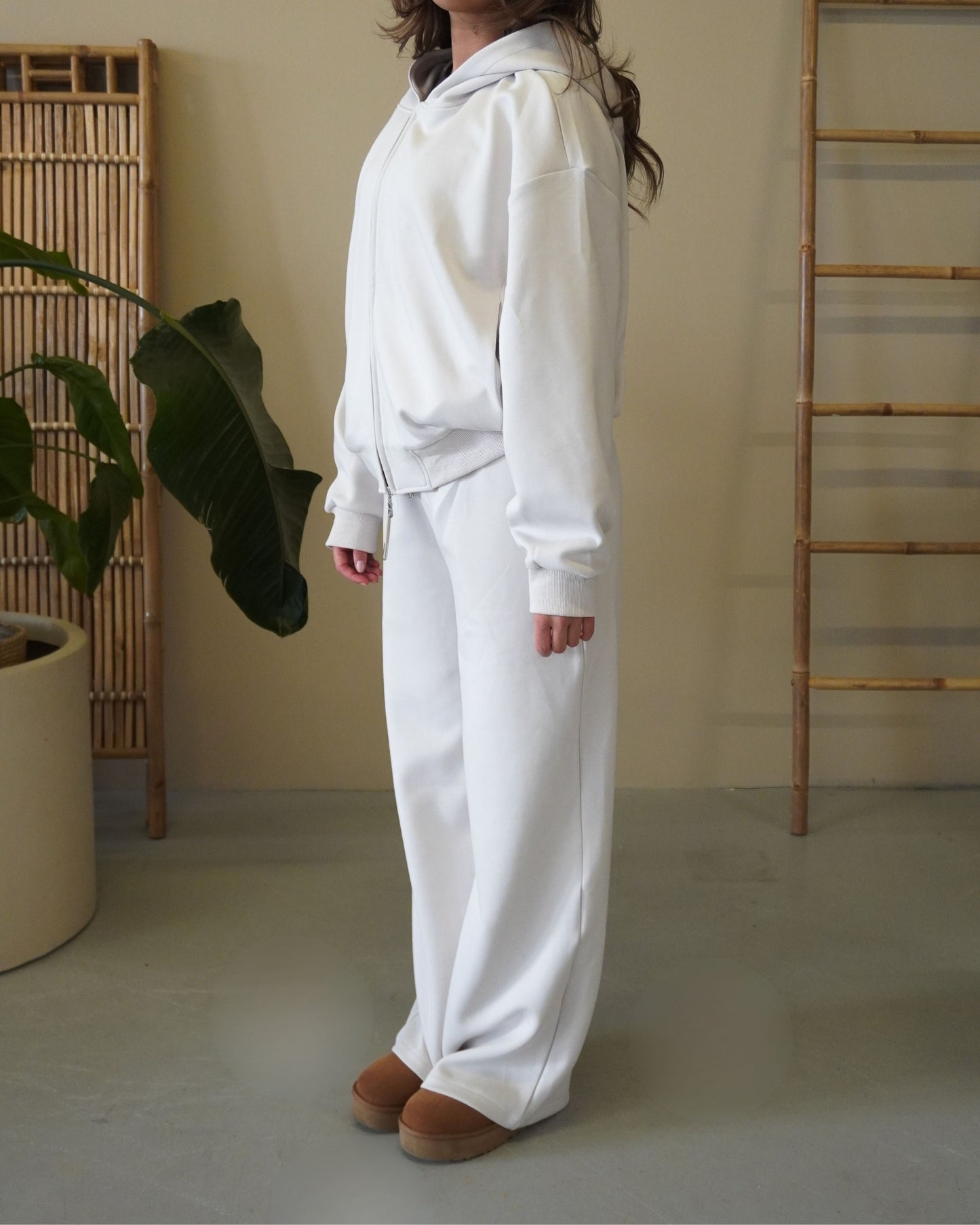 Person wearing a white hoodie and white pants, standing next to a plant.