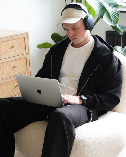 Person sitting on a chair using a laptop, wearing headphones and a cap.