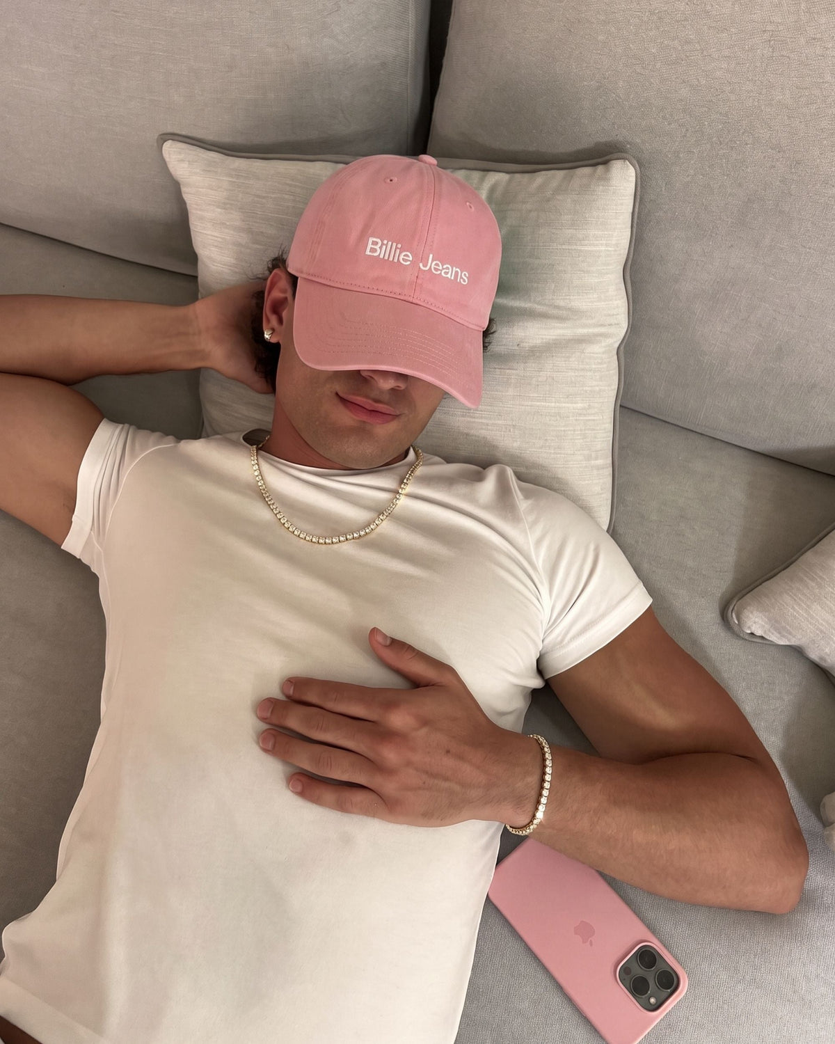 Man in a white T-shirt and pink Billie Jeans cap lying on a sofa wearing the gold Billie Tennis Bracelet 4mm