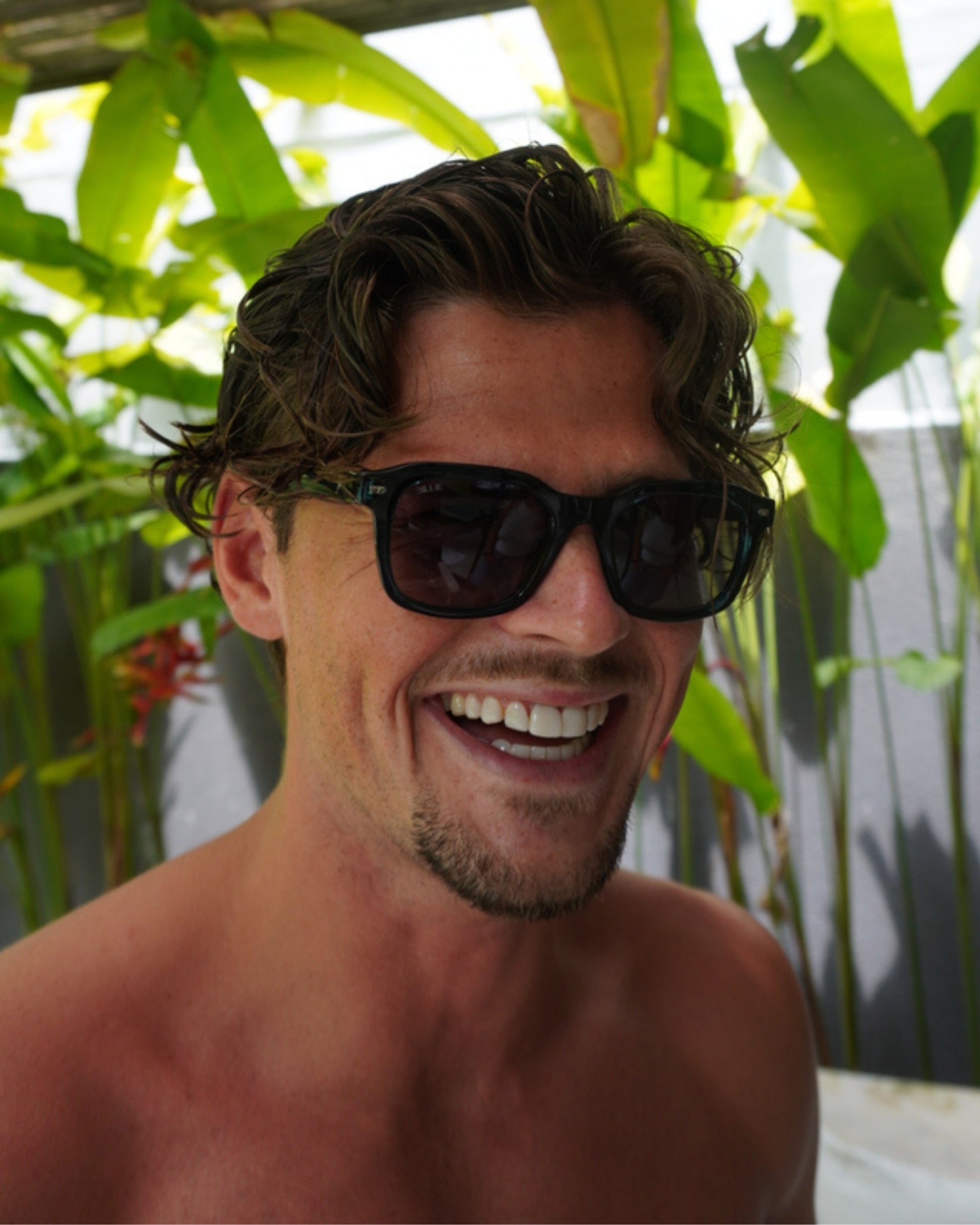 Smiling Person with curly hair wearing sunglasses, surrounded by green plants.
