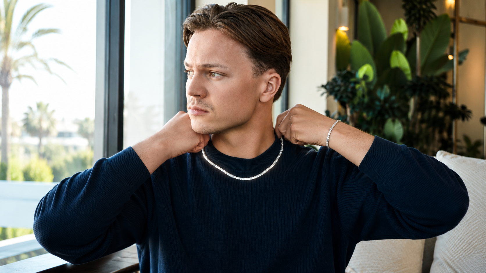 A man wearing a dark navy long-sleeve shirt and a silver beaded necklace, posing indoors near a window with plants in the background.