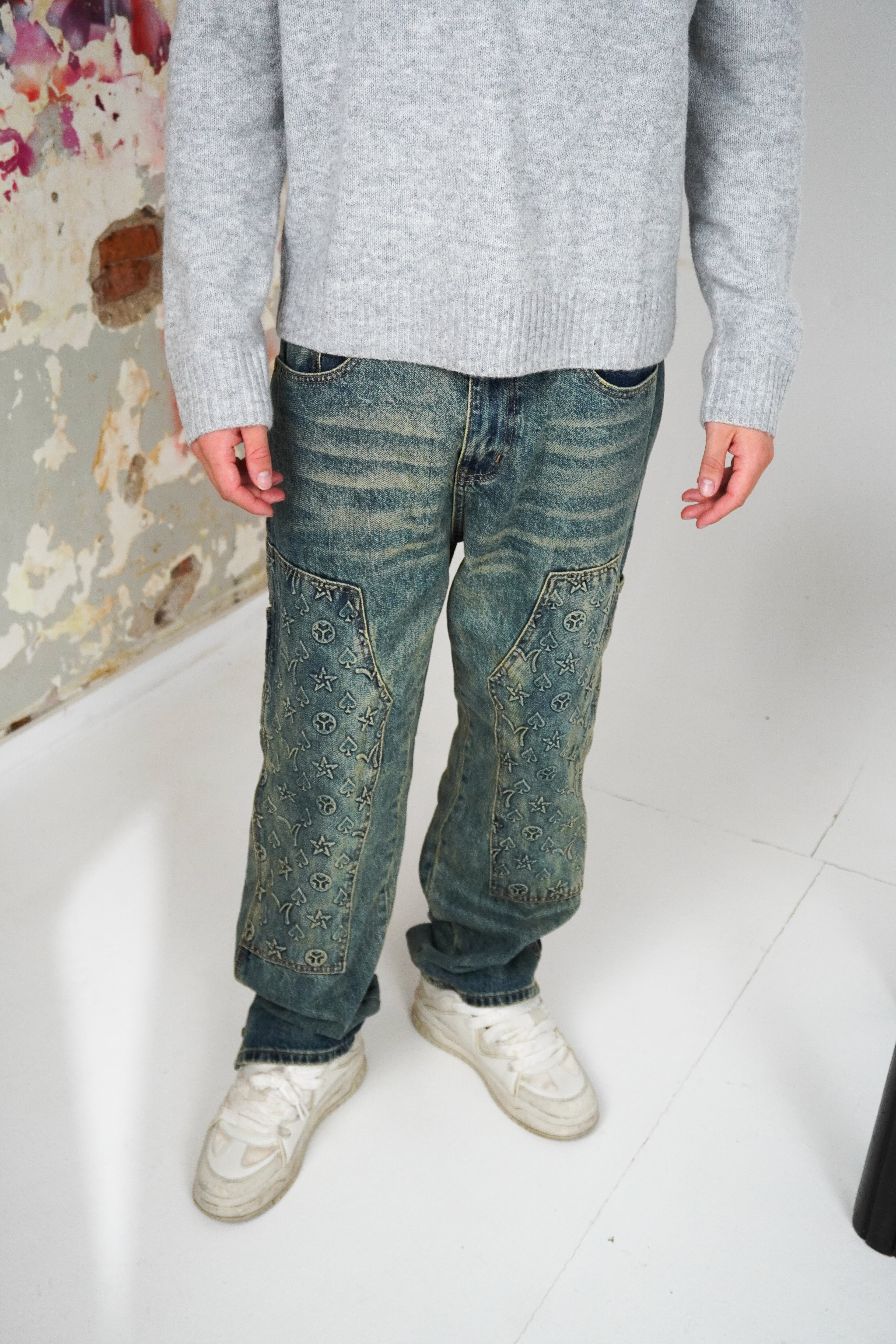 Person wearing a gray sweater, blue jeans, and white shoes on a plain background