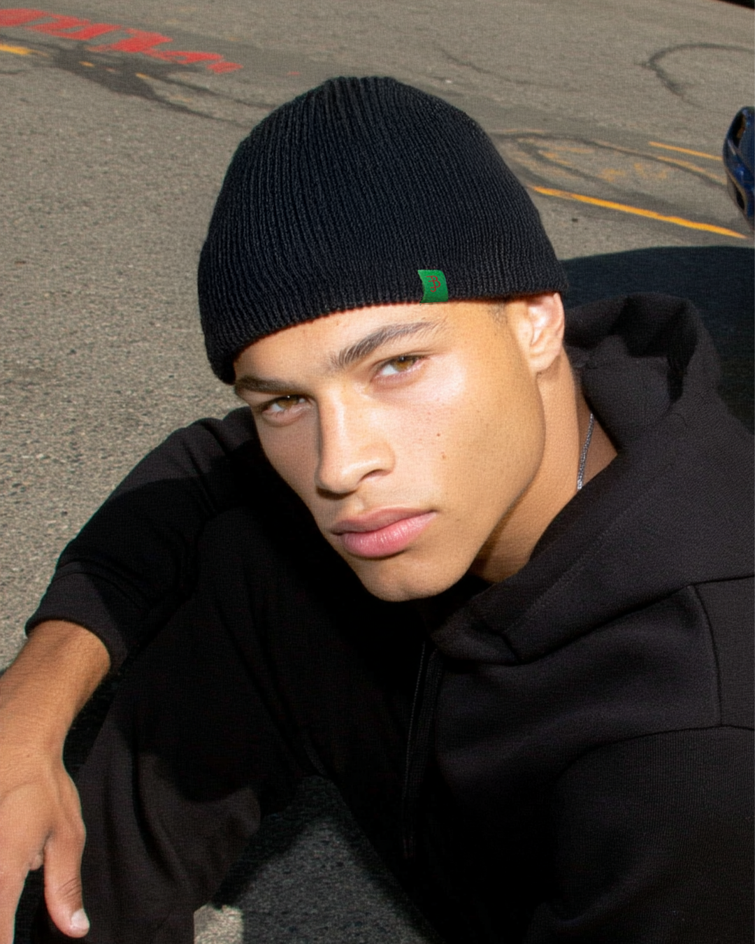 Person wearing a black beanie with a green logo and black hoodie on a street background