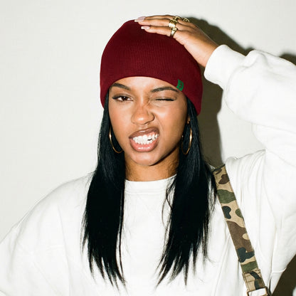 Person wearing a red beanie and white sweatshirt with a camouflage strap over shoulder on a plain background