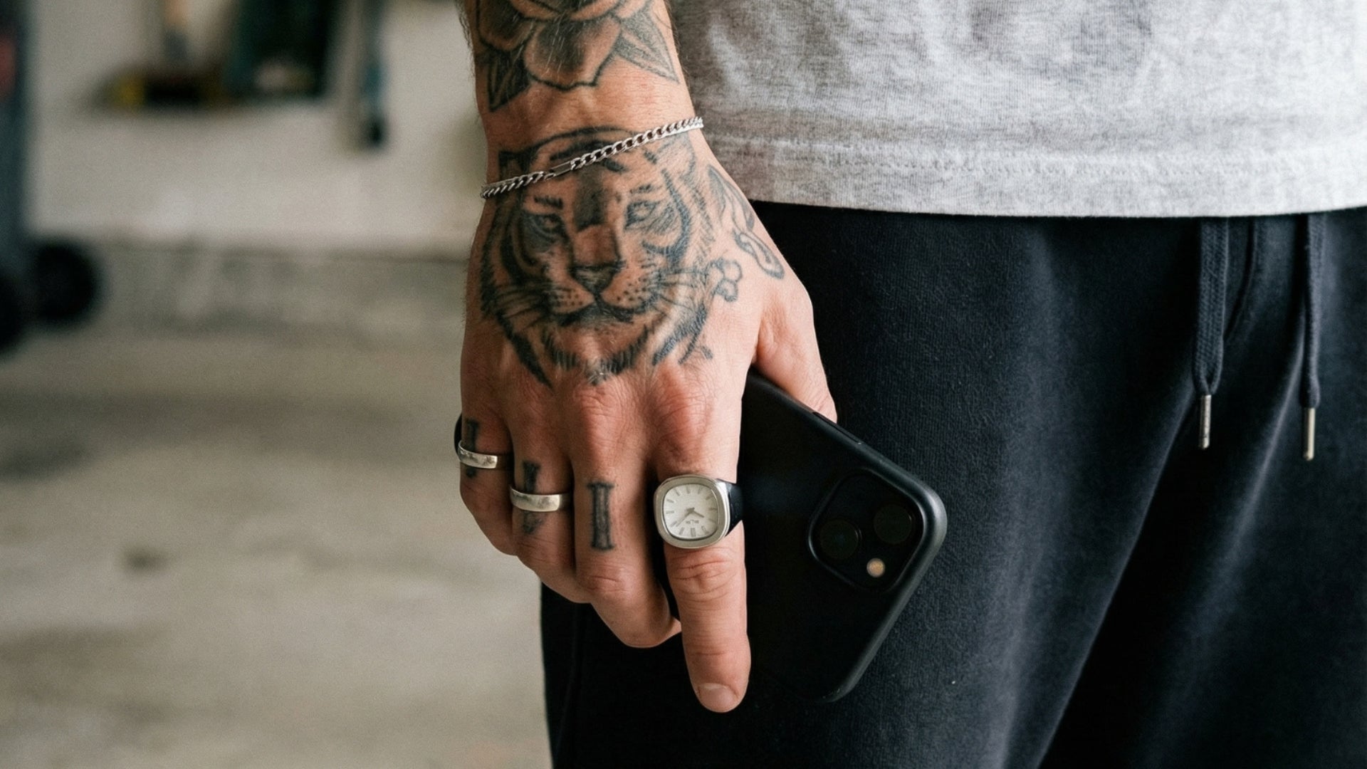A close-up of a tattooed hand holding a black smartphone, adorned with multiple rings and a silver chain bracelet, set against a blurred gym background.