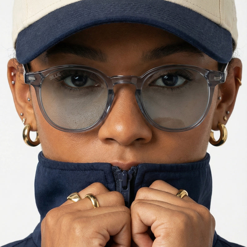 Person wearing glasses, a cap, and a dark jacket with gold hoop earrings and rings.