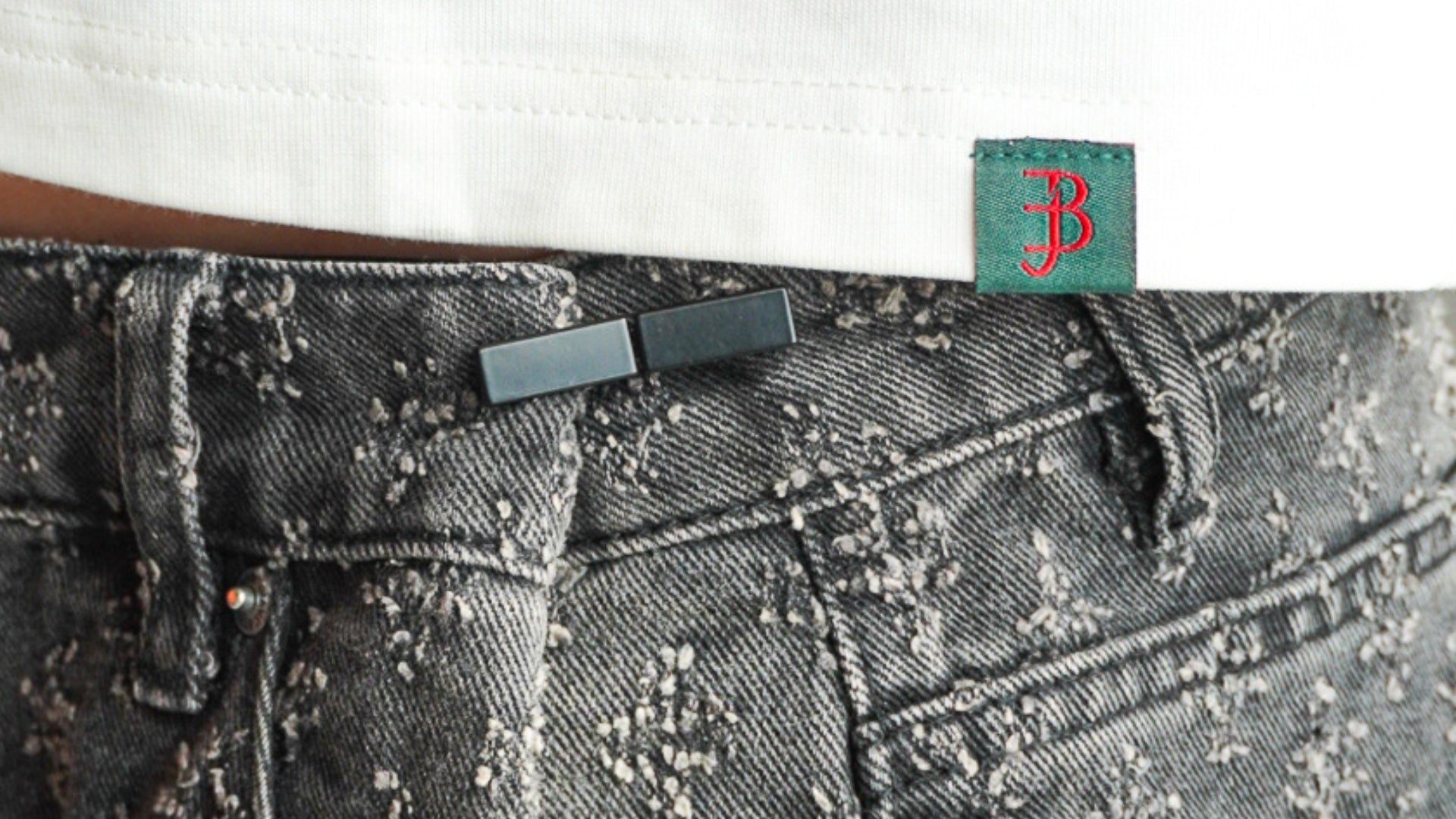 Close-up of a black rectangular belt with a silver-toned buckle worn with distressed dark gray denim jeans and a white shirt. A green fabric tag with a red embroidered 'B' is visible near the waistband.