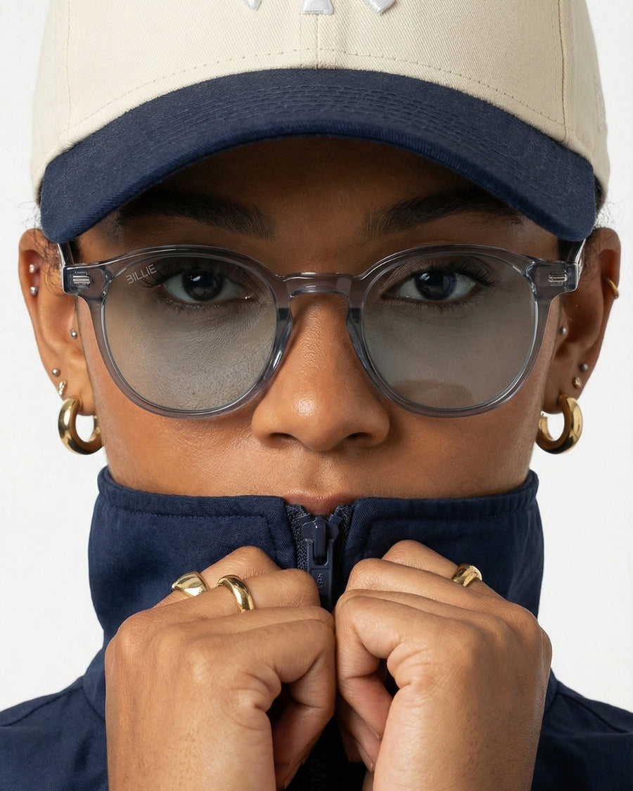 Person wearing a beige cap, sunglasses, and a navy jacket with gold hoop earrings and rings.