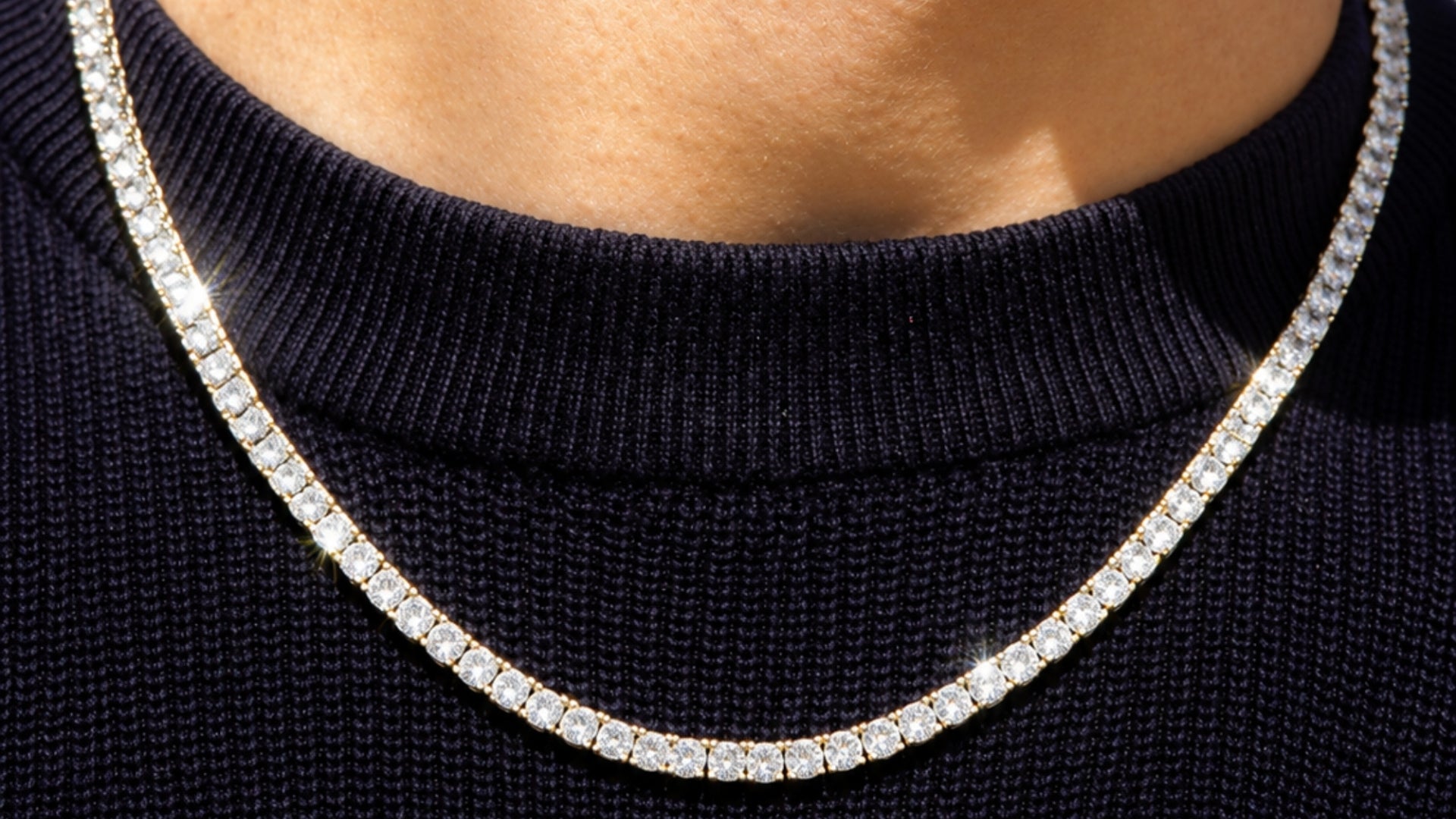 A close-up of a diamond tennis necklace worn around the neck of a person wearing a black ribbed turtleneck sweater.