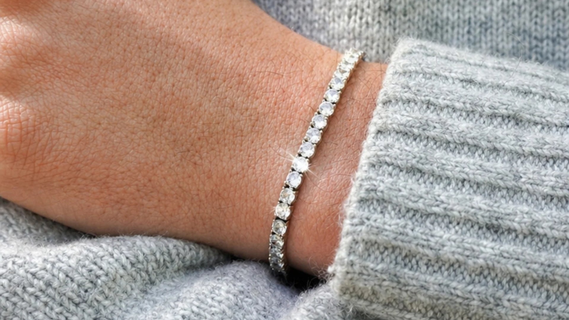 A close-up of a delicate silver tennis bracelet with clear stones worn on a person's wrist, resting against a gray ribbed knit sweater.