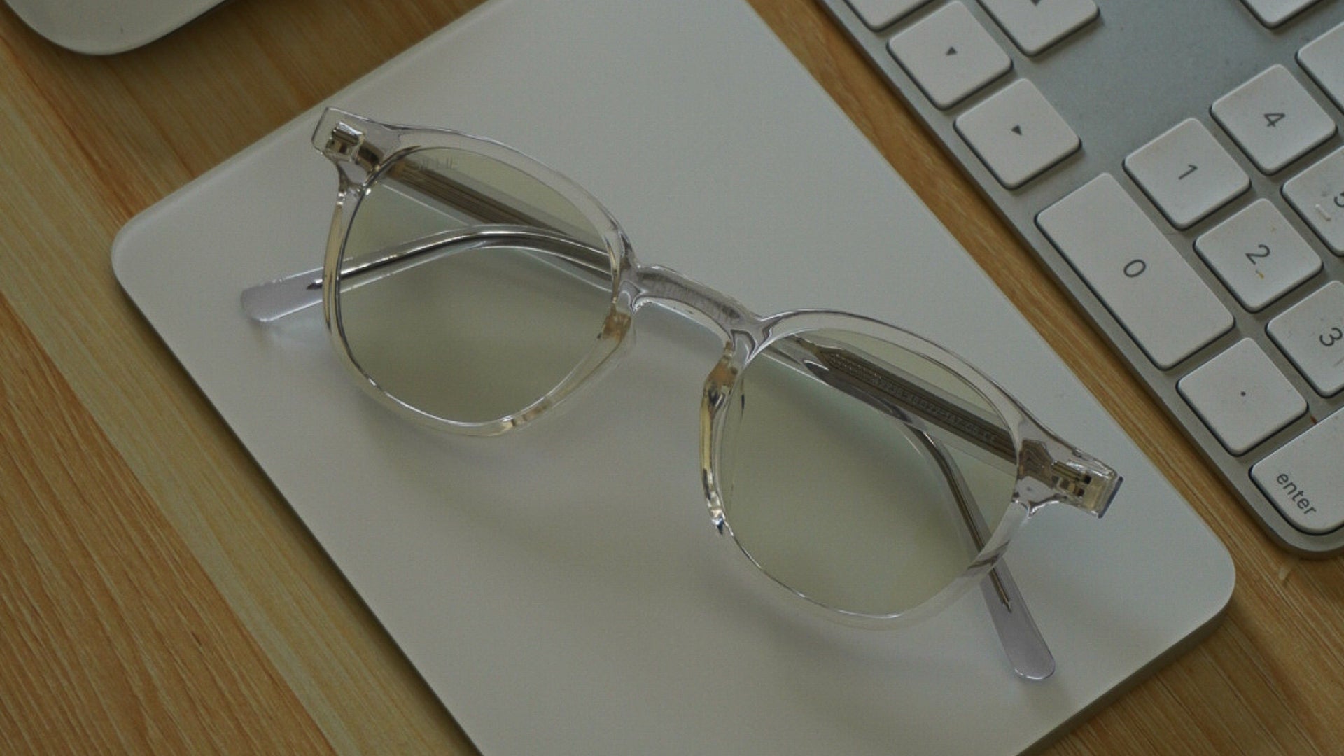 Clear-framed eyeglasses with tinted lenses resting on a white tablet on a wooden desk next to a keyboard.