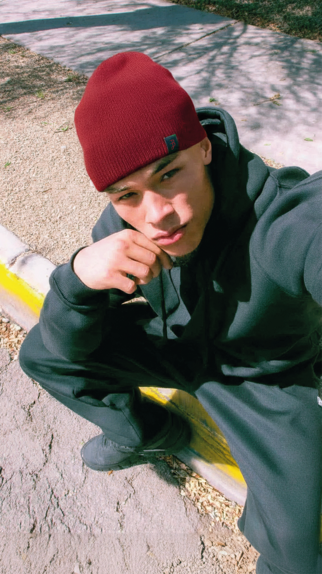 Person wearing a red beanie and dark jacket sitting on a curb.